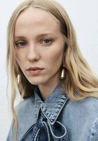 Veste en denim avec un détail nœud papillon, de couleur bleu clair et avec un col texturé. Des boucles d'oreilles pendantes en acier inoxydable rehaussent le look global.