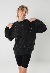 Felpa nera con una vestibilità rilassata, caratterizzata da un motivo a pois bianchi su tutta la superficie e polsini a coste. Indossata con pantaloni shorts neri da bicicletta.