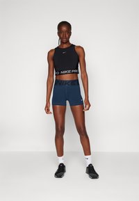 Top corto negro de tirantes con una banda "NIKE PRO", combinado con shorts azul marino que tienen una amplia cintura con el logo "NIKE". El modelo lleva puestas zapatillas deportivas negras.