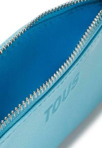 Primer plano de un estuche de cuero azul claro con cremallera, con dientes de cremallera plateados y la palabra "TOUS" grabada en el exterior.