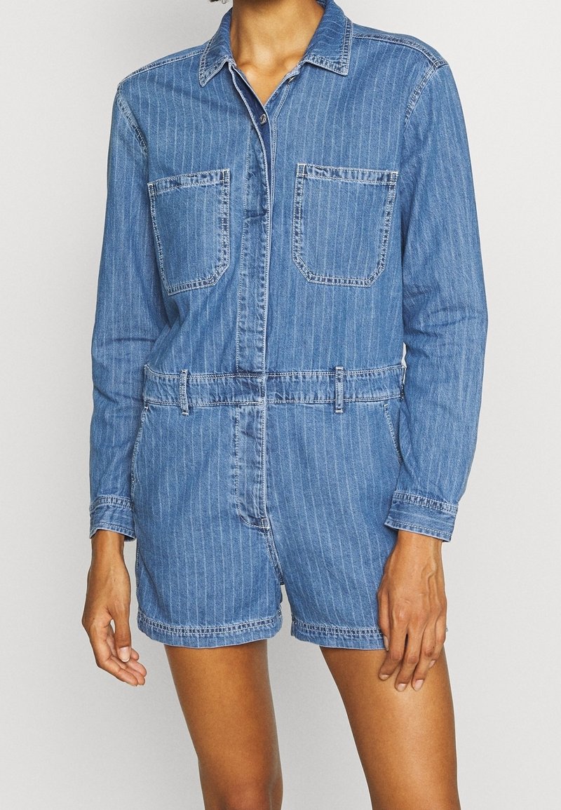 Femme portant une combinaison-short en denim bleu à fines rayures, manches longues, avec des poches avant, debout devant un fond uni.