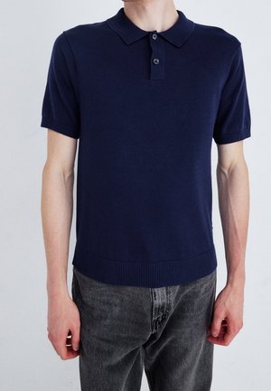 Granatowa koszulka polo z krótkim rękawem, wykonana z dzianiny, z plisą na trzy guziki, prążkowanym kołnierzykiem i dolnym obszyciem, w zestawieniu z szarymi spodniami dżinsowymi.