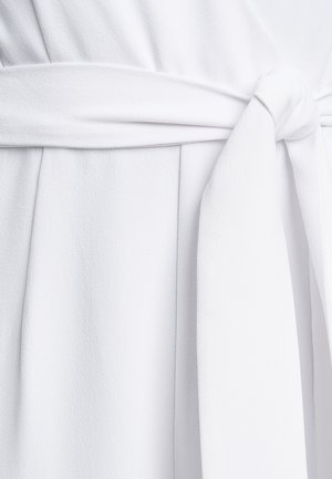 Bijela haljina omotnica s teksturiranom tkaninom, koja ima kravatu u struku i glatku završnicu. Detaljni plisirani detalji poboljšavaju drapiranje i oblik.
