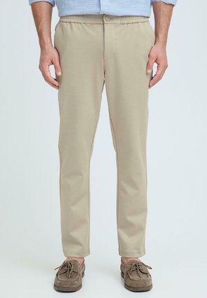 Mężczyzna ubrany w beżowe casualowe spodnie z elastycznym pasem i guzikiem, w połączeniu z brązowymi zamszowymi mokasynami, stojący na jednolitym tle.