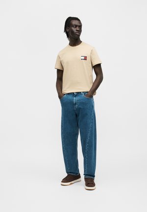 Man met handen in de zakken, draagt een beige T-shirt met korte mouwen en logo, losse blauwe jeans en bruine sneakers, kijkt iets naar rechts.