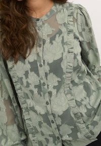 Light green blouse featuring a textured floral pattern, frill detailing along the button placket, and long sleeves with gathered accents.