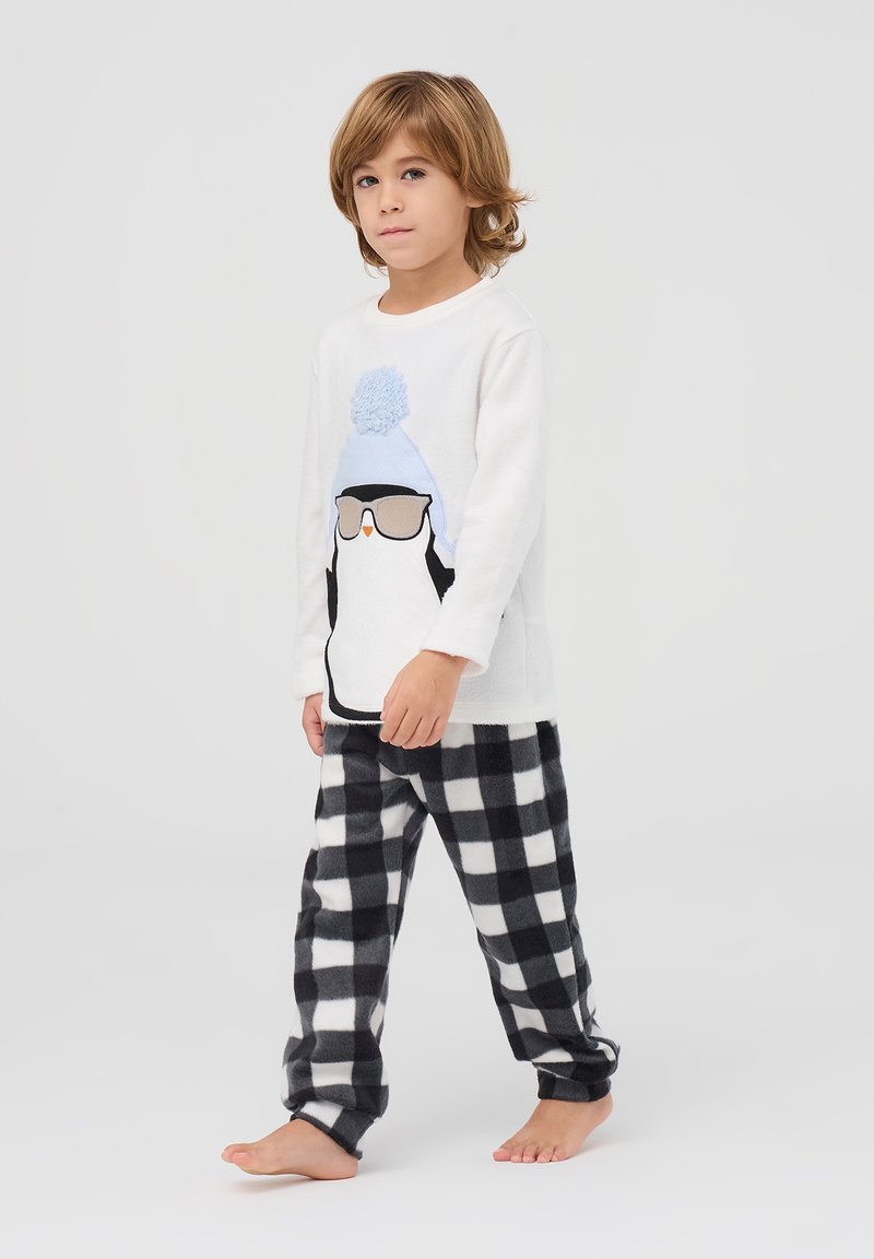 Biała bluzka z długim rękawem z graficznym motywem pingwina i niebieską czapką z pomponem, w połączeniu z czarno-białymi spodniami z polarowego materiału w kratkę.