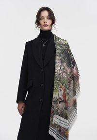 Écharpe en laine présentant une scène de renard et de nature dans des couleurs vives, drapée sur un manteau noir, avec une frange délicate et une bordure rayée.