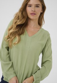 Blouse vert clair, texturée, avec un décolleté en V et des manches longues légèrement bouffantes. Présente une fente subtile au niveau du décolleté et un tissu doux.