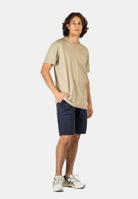 Tan cotton t-shirt with a crew neckline, paired with navy shorts. Model wears white sneakers with a light blue accent.