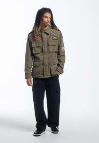 Militarystyczna oliwkowa kurtka z wieloma kieszeniami i naszywkami, zestawiona z luźnymi czarnymi spodniami cargo i czarnymi trampkami.
