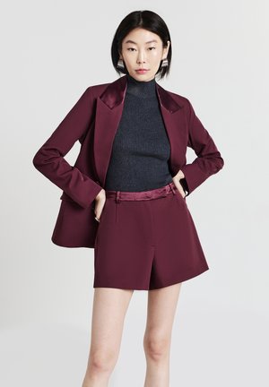 Femme portant un blazer bordeaux et un short assorti avec un pull à col roulé noir à côtes, se tenant les mains sur les hanches devant un fond uni.