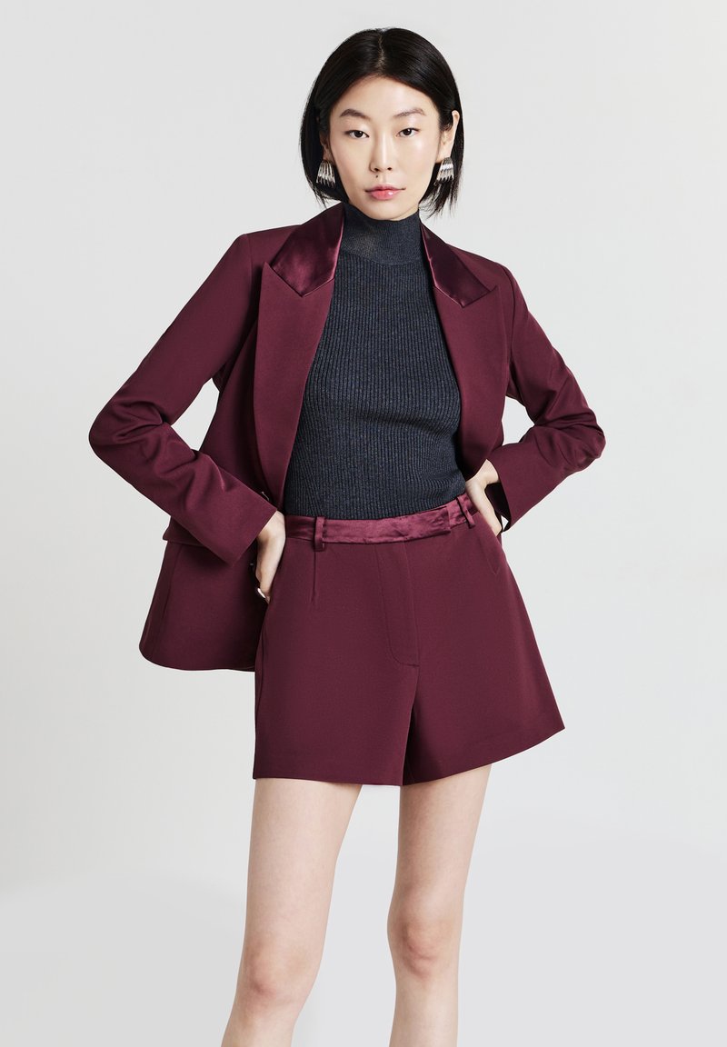 Donna che indossa un blazer bordeaux e pantaloni corti abbinati, con un maglione a collo alto nero a coste, in piedi con le mani sui fianchi davanti a uno sfondo semplice.