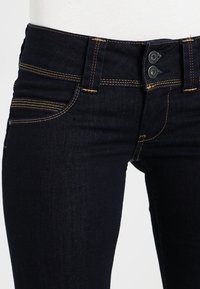 Mörkblå denimjeans med kontrasterande gul sömnad, dubbel knappstängning och framfickor på vit bakgrund.