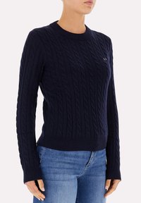 Maglione blu navy a treccia con scollatura a girocollo e polsini a costine. Presenta una silhouette aderente e un sottile dettaglio del logo sul petto.