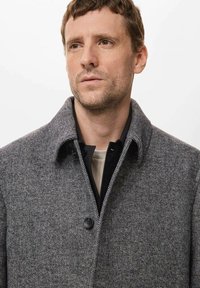 Abrigo de lana en espiga gris con un gran cuello, cierre de un solo botón y superpuesto a una camisa negra, con una textura suave.