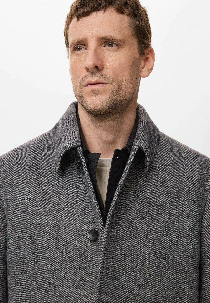 Grå herringbone ullkappa med stor krage, enfaldig knappstängning och lagd över en svart skjorta med en mjuk textur.