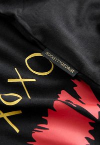 Must mustriga mustriga kuldse ja punase trükitud kujundusega. Sisaldab teksti ja abstraktseid kujundeid. Sildil on kirjutatud "ROCKETT ST. GEORGE" mustal ristkülikulisel sildil.