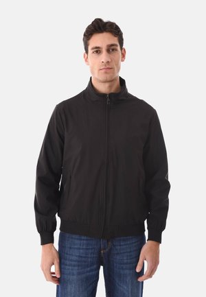 Uomo che indossa una giacca nera con cerniera e collo alto e jeans blu, in piedi contro uno sfondo bianco semplice.