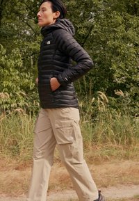 Giacca a vento nera con cappuccio, con chiusura a zip anteriore e tasche laterali. Pantaloni cargo khaki con tasche laterali, indossati con scarpe scure.