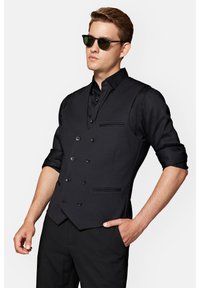 Czarna kamizelka dwurzędowa o teksturowanym wykończeniu, stylizowana na czarnej koszuli. Charakteryzuje się eleganckim krojem, dopasowanym fasonem i subtelnymi akcentami.