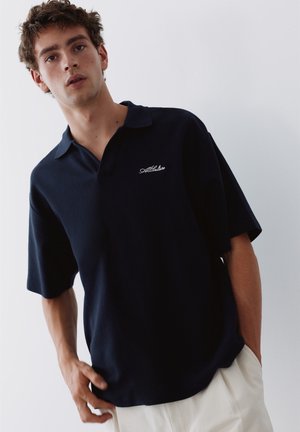 Jeune homme aux cheveux bouclés portant un polo bleu marine foncé à manches courtes et un pantalon clair sur un fond uni.