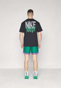 Camiseta negra de manga corta con un gran logo "NIKE" en gris y verde, combinada con pantalones cortos verdes con ribetes morados y zapatillas verdes.