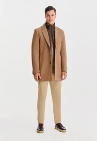 Wełniany płaszcz w kolorze tan z subtelną teksturą, nałożony na ciemną pikowaną kurtkę. Kremowe spodnie i brązowe sneakersy dopełniają strój.