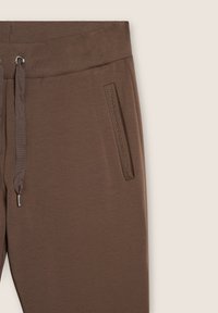 Pantaloni joggers marroni con vita regolabile, coulisse e tasche con zip. Il materiale ha una consistenza liscia e un design aderente.