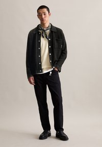 Donkergroene jas met witte knopen over een lichtgele trui, zwarte broek en zwarte schoenen. Een gestructureerde sjaal is om de hals geknoopt.