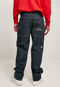 Dark wash denim pants feature multiple pockets, contrasting stitching, and branding on the waistband and back pocket. Worn with a red top.
