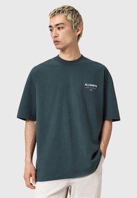 Jeune homme aux cheveux blonds courts portant un t-shirt surdimensionné teal foncé avec un texte blanc sur la poitrine et un pantalon beige clair, les mains dans les poches.