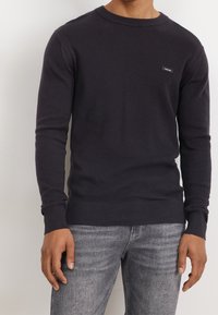 Maglione blu navy a maniche lunghe con superficie testurizzata, scollo a girocollo e un piccolo badge nero sul petto. Indossato con jeans grigi.