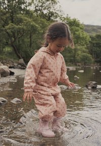 Roze regenpak met bloemenpatroon, voorzien van een capuchon, elastische boorden en ritssluiting, gecombineerd met lichtroze waterdichte laarzen, spetterend in het water.