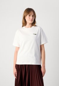Hvit bomull T-skjorte med korte ermer, prydet med en grønn brodert logo på brystet. Avslappet passform og glatt tekstur.