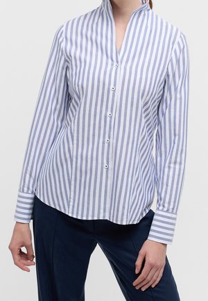 Donna che indossa una camicia bianca a maniche lunghe con righe verticali blu e pantaloni blu scuro, in piedi con una mano vicino all'anca.