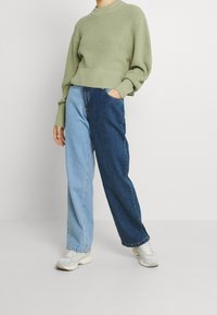 Pull vert clair en maille à col rond, associé à un jean ample bicolore (denim clair et foncé). Des baskets blanches complètent la tenue.