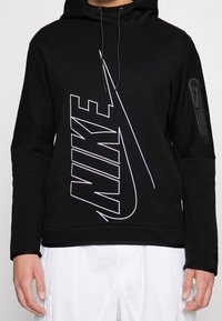 Sudadera negra con capucha, que presenta un gran logo blanco de Nike, cordones en la capucha y un bolsillo con cremallera en la manga izquierda. Fabricada en un material suave y texturizado.