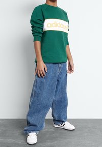 Grön sweatshirt med vitt och gult "adidas"-logotyp, kombinerad med lösa blå denimjeans och vita sneakers med svarta accenter.