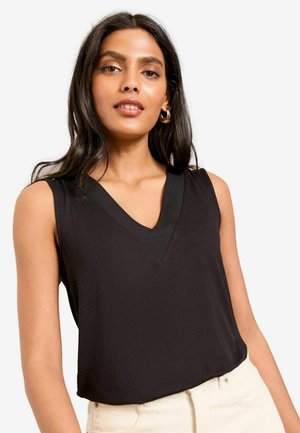 Pikkade tumedate juustega naine, kes kannab kuldseid rõngaskõrvarõngaid, musta varrukateta V-kaelusega särki ja heledaid pükse, valge taustaga.