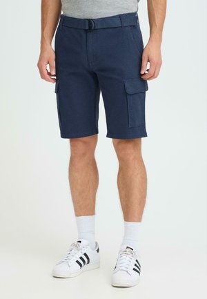 Mann trägt marineblaue Cargoshorts mit Gürtel, weiße Socken und weiße Turnschuhe mit schwarzen Streifen, steht vor einem schlichten Hintergrund.