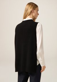 Czarny dziergany sweter bez rękawów z okrągłym dekoltem, teksturowaną tkaniną i luźnym krojem, noszony na białej koszuli z kołnierzykiem.