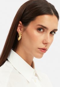 Guldörhängen med en fläktliknande design, med ribbade detaljer. Modellen bär en vit skjorta med krage, som visar upp örhängena.