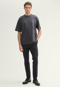 Dunkelgraues Kurzarm-T-Shirt mit geradem Schnitt und einem vertikalen weißen Logo-Druck, kombiniert mit dunklen, taillierten Hosen und schwarzen Schuhen.