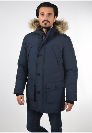 Tamno plava puffer jakna s kapuljačom obloženom krznom, koja ima zatvaranje na patentni zatvarač i gumb, četiri prednja džepa i prilagođeni kroj s rebrastim manžetnama.