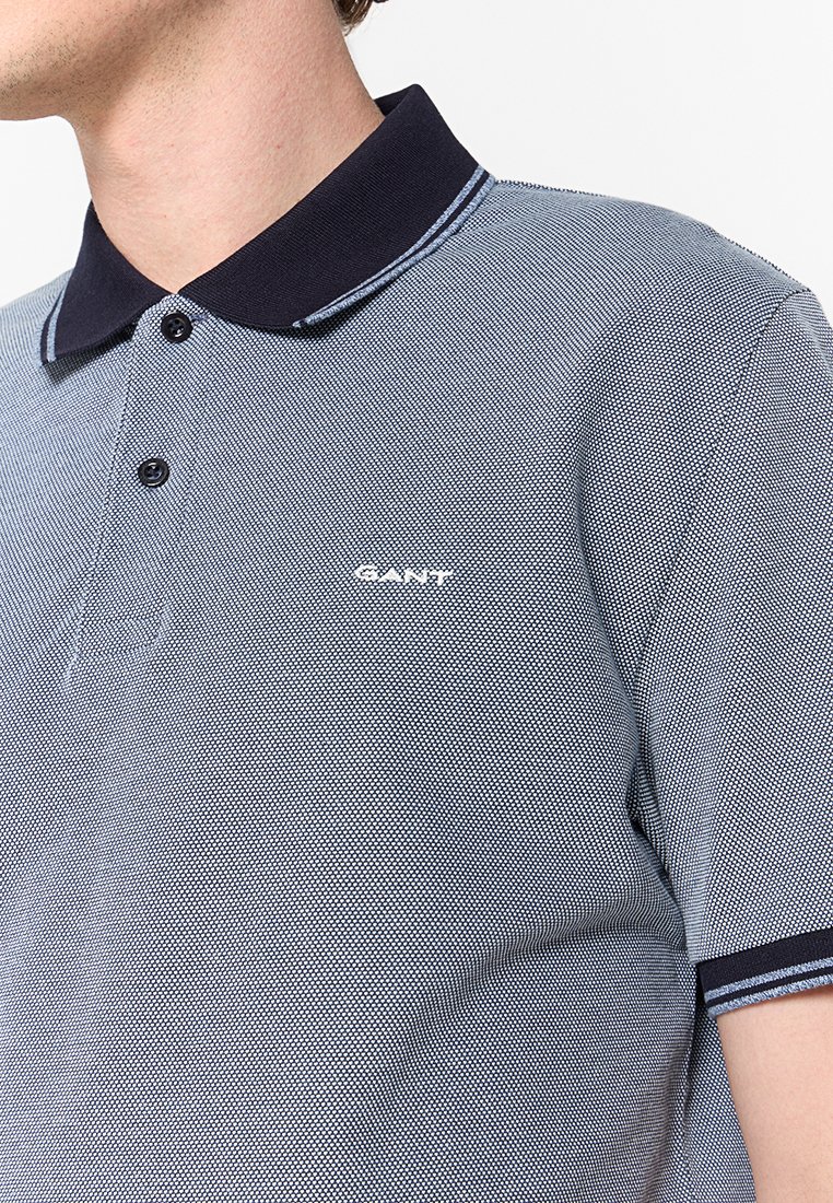 Polo shirt met korte mouwen in lichtblauw met een gestructureerd patroon, marineblauwe kraag en wit logo op de borst. Beschikt over twee knopen en gestreepte accenten.