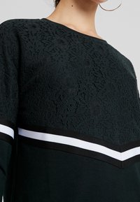 Felpa verde scuro con una parte superiore in pizzo, caratterizzata da un motivo a zigzag e un bordo nero con strisce bianche all'orlo e alle maniche.