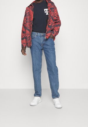 Rot-schwarz gemusterte Zip-Jacke über einem marineblauen T-Shirt, kombiniert mit hellblauen Jeans und weißen Sneakers. Lässiges Outfit, entspannte Passform.