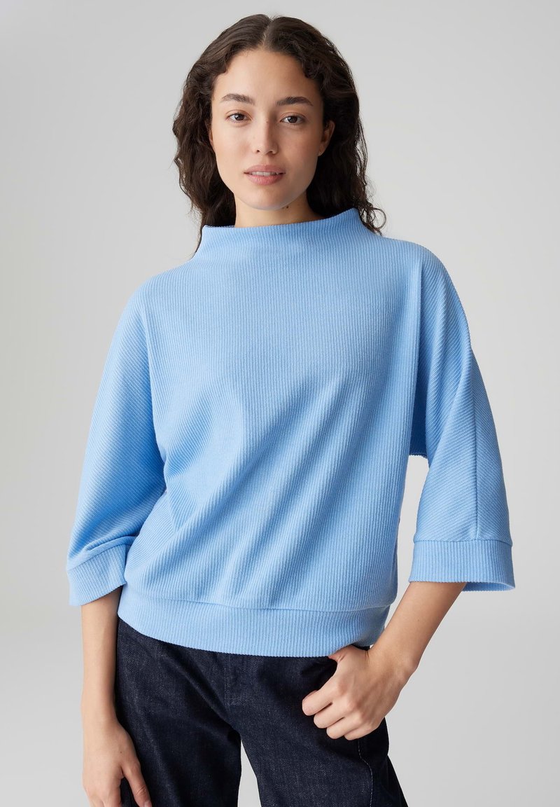 Hellblauer Pullover aus geripptem Stoff, mit hohem Kragen, dreiviertellangen Ärmeln und lockerer Passform. Kombiniert mit dunklen Jeans.