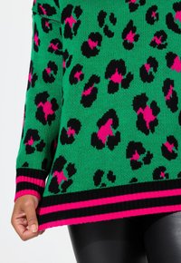 Grüner gestrickter Pullover mit einem Leopardenmuster in Schwarz und Rosa, mit einem schwarz-rosa gestreiften Saum. Die Textur wirkt weich und gemütlich.
